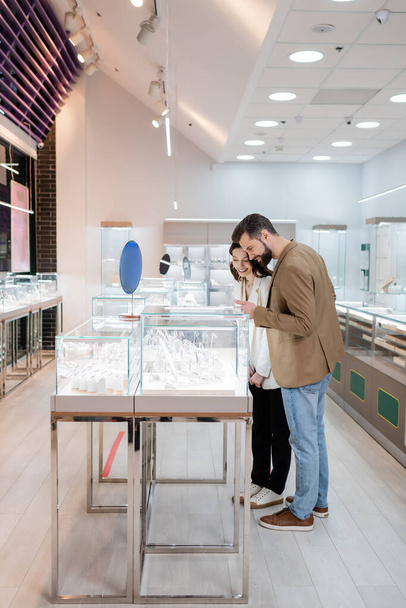 Klient wskazujący na biżuterię w pobliżu uśmiechniętej dziewczyny w sklepie  - Zdjęcie, obraz