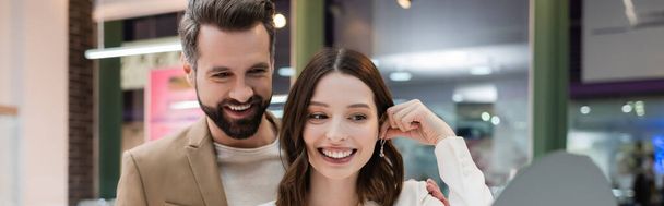 Smiling woman holding earring near mirror and boyfriend in jewelry store, banner  - Photo, Image
