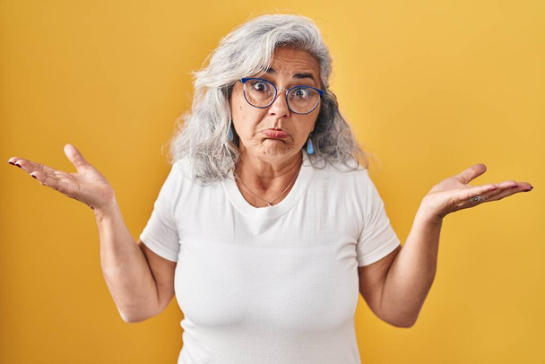 Femme d'âge moyen avec les cheveux gris debout sur fond jaune expression inutile et confus avec les bras et les mains levées. concept de doute.  - Photo, image