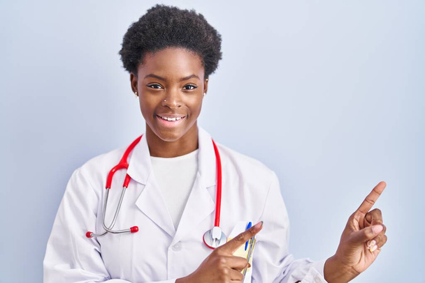 African american woman wearing doctor uniform and stethoscope smiling and looking at the camera pointing with two hands and fingers to the side.  - Foto, Imagen