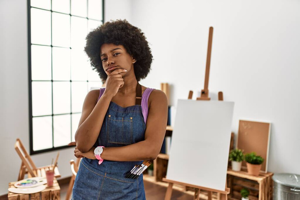 Joven mujer afroamericana con cabello afro en el estudio de arte mirando confiado a la cámara sonriendo con los brazos cruzados y la mano levantada en la barbilla. pensar positivamente.  - Foto, Imagen