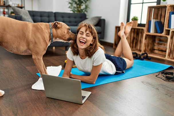 Νεαρή καυκάσια γυναίκα αθλήτρια χαμογελώντας αυτοπεποίθηση χρησιμοποιώντας φορητό υπολογιστή με το σκυλί στο σπίτι - Φωτογραφία, εικόνα