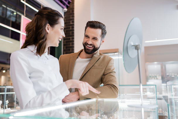 Positive customer looking at young seller pointing at showcase in jewelry shop - Photo, Image