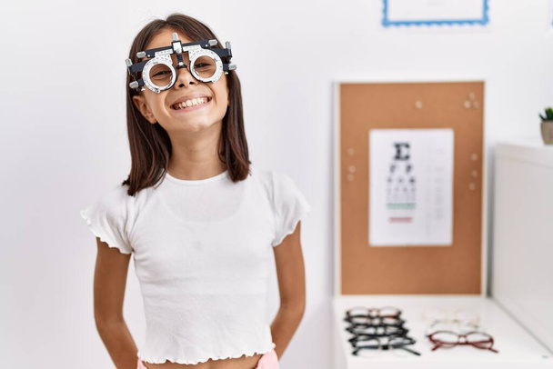 Молода іспаномовна дівчина в окулярах з оптометрією виглядає позитивно і щасливо стоячи і посміхаючись з впевненою посмішкою, показуючи зуби
  - Фото, зображення