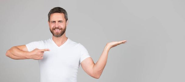 Happy handsome man smile pointing finger at open hand grey background copy space, presenting product. Man face portrait, banner with copy space - Photo, image