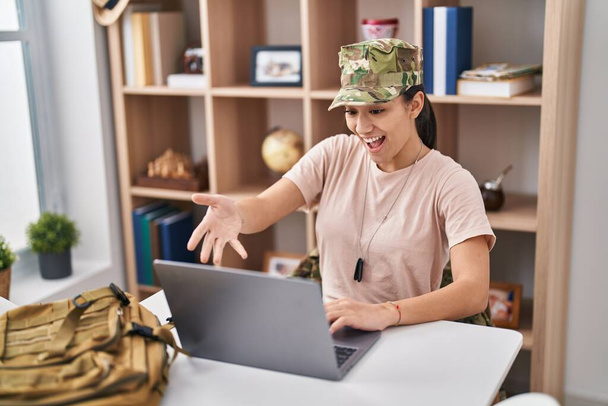 Young south asian woman wearing camouflage army uniform doing video call celebrating achievement with happy smile and winner expression with raised hand  - Foto, Imagen