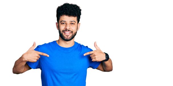 Junger arabischer Mann mit Bart trägt legeres blaues T-Shirt und sieht selbstbewusst aus mit einem Lächeln im Gesicht, zeigt stolz und glücklich mit den Fingern.  - Foto, Bild