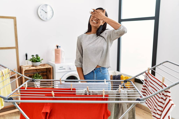 Young hispanic woman putting fresh laundry on clothesline smiling and laughing with hand on face covering eyes for surprise. blind concept.  - Photo, Image
