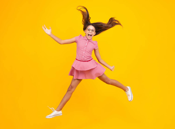 Amazed teen girl. Excited expression, cheerful and glad. Full length cheerful teenager kid jump enjoy rejoice win isolated on yellow background. Small child girl in summer dress jumping - Zdjęcie, obraz