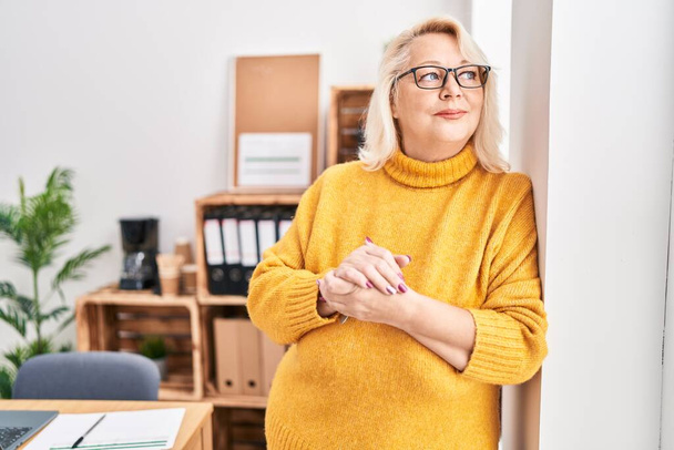 Μέση ηλικία ξανθιά γυναίκα επιχειρηματίας χαμογελά αυτοπεποίθηση στέκεται στο γραφείο - Φωτογραφία, εικόνα