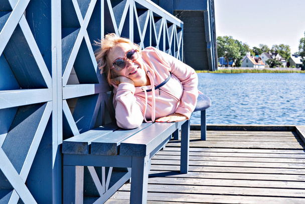 Frau mittleren Alters in rosa Kapuzenpulli lehnt auf einer Pantone-Bank am Seeufer, lächelt und blickt in die Kamera - Foto, Bild