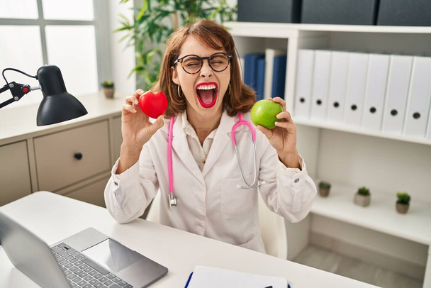 Young doctor woman holding heart and green apple smiling and laughing hard out loud because funny crazy joke.  - 写真・画像