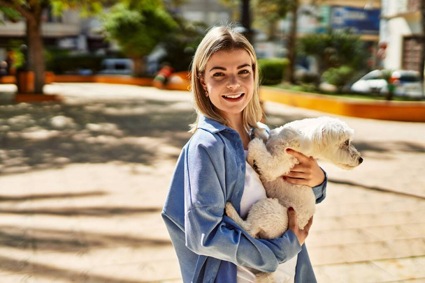 Young blonde girl smiling happy holding dog at the city. - Photo, Image