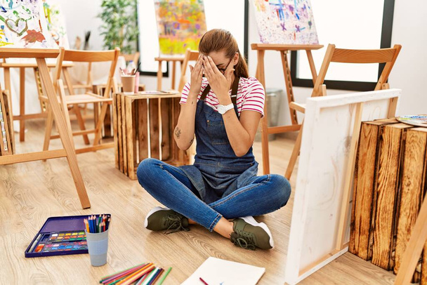 Молодая брюнетка в художественной студии сидит на полу с грустным лицом, прикрывающим лицо руками во время плача. депрессия.  - Фото, изображение