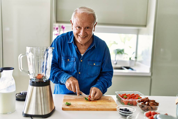 Senior man smiling confident cutting strawberry at kitchen - Фото, изображение