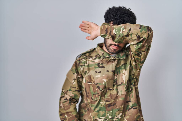 Арабский мужчина в камуфляжной армейской форме закрывает глаза рукой, выглядит серьезным и грустным. беспросветная, скрывающая и отвергающая концепция  - Фото, изображение