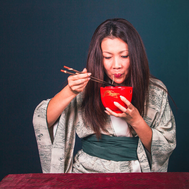 Linda mulher asiática bonito comer macarrão asiático com pauzinhos de madeira. estúdio filmado em um fundo verde escuro - Foto, Imagem