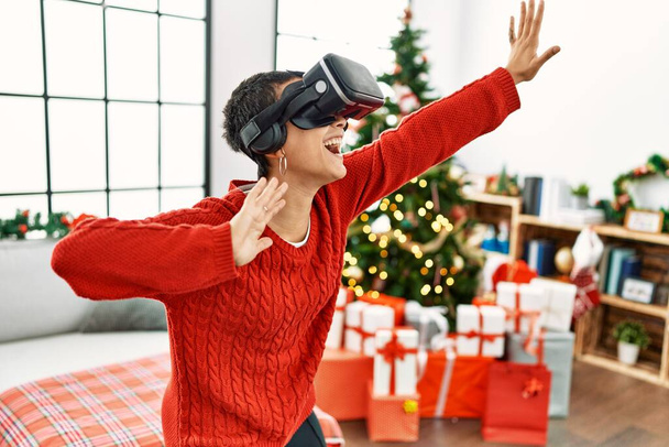 Young hispanic woman playing video game using vr glasses standing by christmas tree at home - Photo, Image