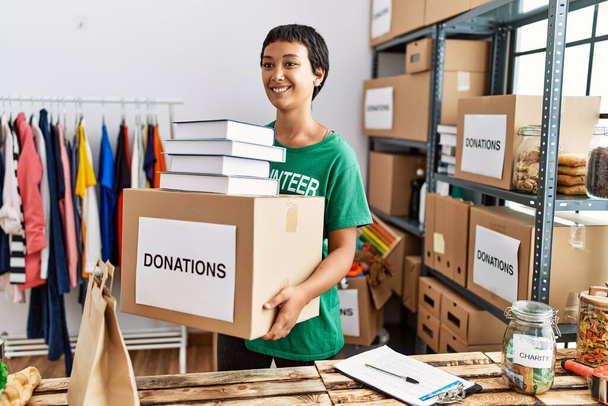 Mladá hispánka v dobrovolnické uniformě, držící v charitativním centru balíček dárcovství knih - Fotografie, Obrázek