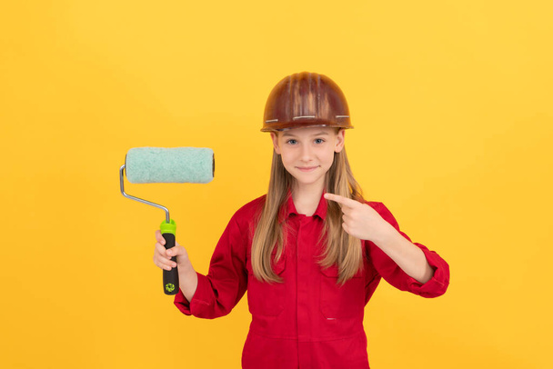 happy teen child in builder hard hat with paint roller on yellow wall. pointing finger. - Foto, Bild