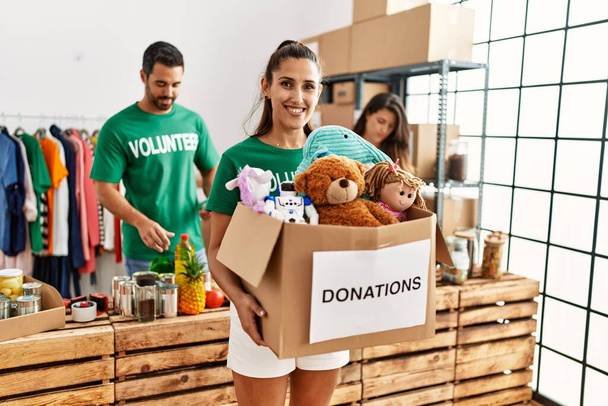 Group of hispanic volunteers working at charity center. Woman smiling happy holding box with toys to donate. - Photo, Image