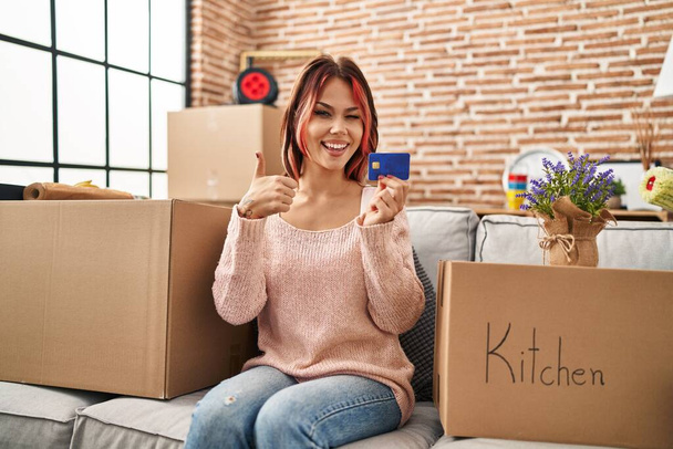 Молода біла жінка переїжджає в новий будинок, тримає кредитну картку посміхаючись щасливо і позитивно, великий палець робить відмінний знак схвалення
  - Фото, зображення