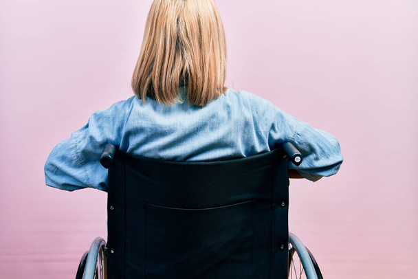 Beautiful blonde woman sitting on wheelchair standing backwards looking away with arms on body  - Photo, Image