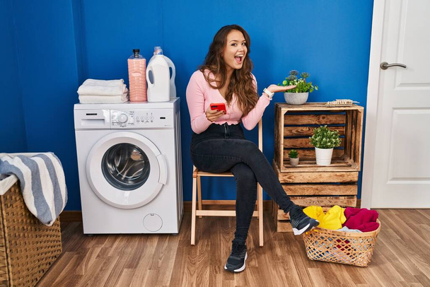 Mujer hispana joven sentada esperando la lavandería usando un teléfono inteligente apuntando a un lado con las manos abiertas mostrando espacio de copia, presentando anuncio sonriendo emocionada feliz  - Foto, imagen