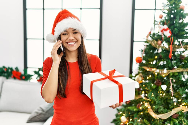 Young latin woman talking on the smartphone standing by christmas tree at home - Photo, Image