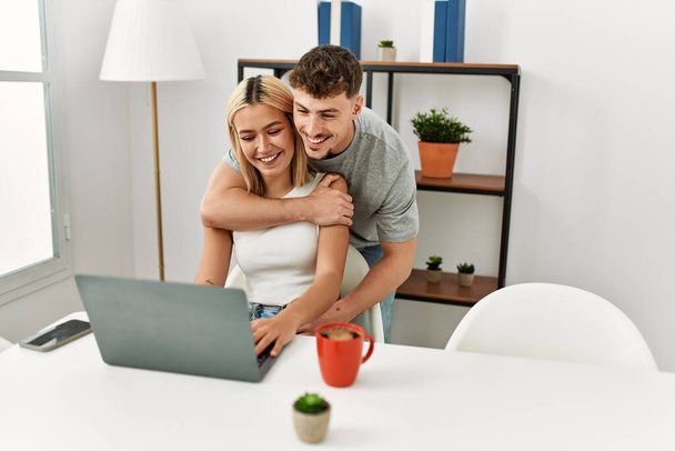 Young caucasian couple hugging and using laptop sitting on the desk at home. - Photo, Image
