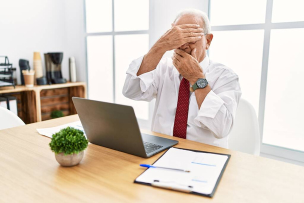 Старший чоловік працює в офісі, використовуючи комп'ютерний ноутбук, що закриває очі і рот руками, здивований і шокований. приховування емоцій
  - Фото, зображення