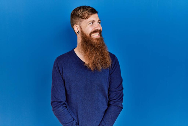 Рудий чоловік з довгою бородою носить випадковий синій светр на синьому фоні, дивлячись на бік з посмішкою на обличчі, природним виразом. впевнений сміх
.  - Фото, зображення