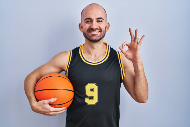 Jeune homme chauve avec barbe portant un uniforme de basket-ball tenant le ballon souriant positif faisant signe ok avec la main et les doigts. expression réussie.  - Photo, image