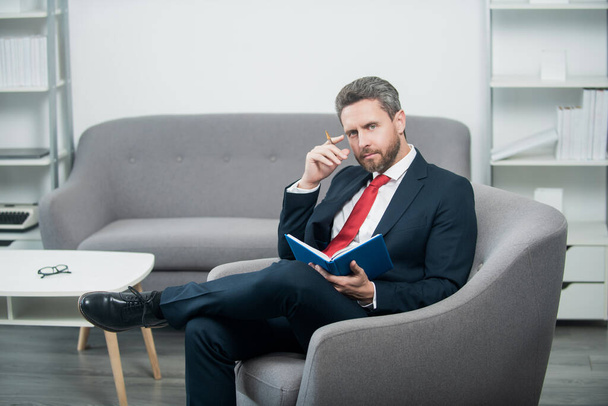 pondering mature businessman in suit sit in office making notes in notebook. - 写真・画像