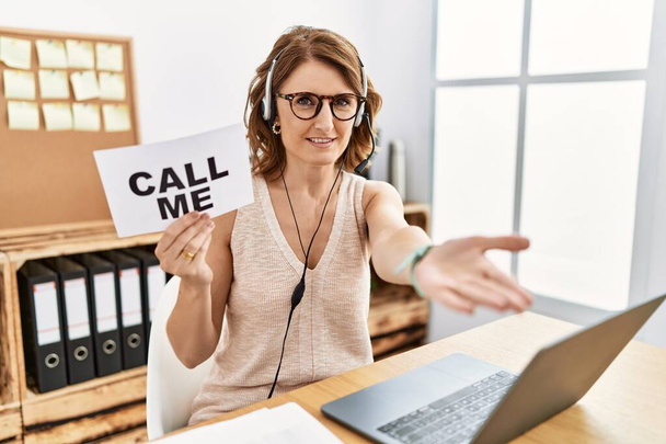 Middle age brunette woman wearing operator headset holding call me banner smiling cheerful offering palm hand giving assistance and acceptance.  - Foto, afbeelding