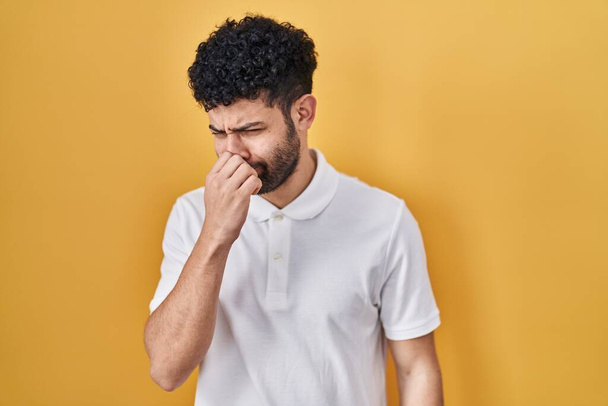 Arabische man die over gele achtergrond staat en iets stinkt en walgelijk ruikt, een ondraaglijke geur, adem inhouden met vingers op de neus. slechte geur  - Foto, afbeelding