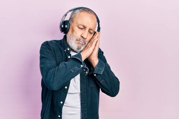 Handsome senior man with beard listening to music using headphones sleeping tired dreaming and posing with hands together while smiling with closed eyes.  - Foto, immagini