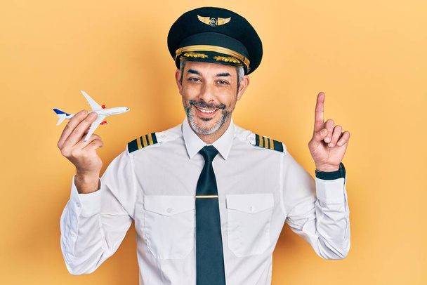Hombre guapo de mediana edad con pelo gris llevando uniforme de piloto de avión sosteniendo avión de juguete sorprendido con una idea o pregunta apuntando con el dedo con la cara feliz, número uno  - Foto, Imagen