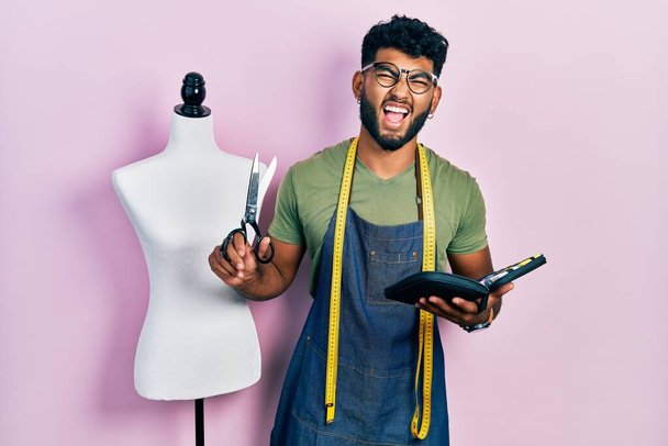 Arab man with beard dressmaker designer holding scissors and sewing kit smiling and laughing hard out loud because funny crazy joke.  - Fotoğraf, Görsel