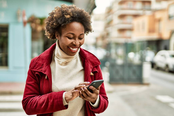 Красивая деловая африканская американка с афроволосами улыбается счастливой и уверенной в себе на открытом воздухе в городе, используя смартфон - Фото, изображение