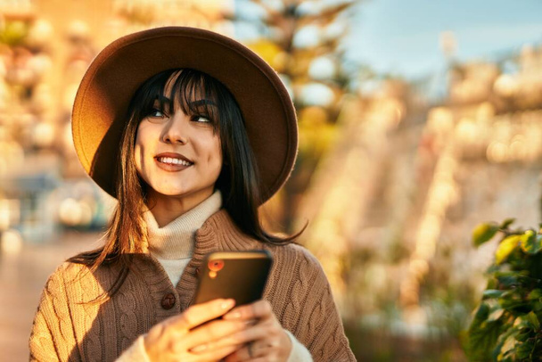 Mujer morena con sombrero de invierno sonriendo usando smartphone en el parque - Foto, imagen