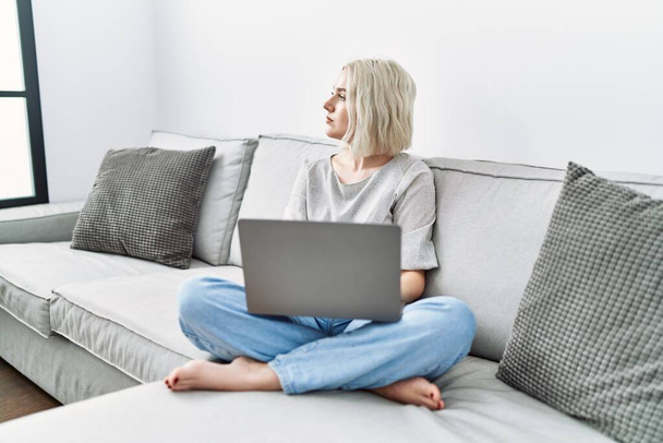 Nuori valkoihoinen nainen, joka käyttää kannettavaa tietokonetta kotona istumassa sohvalla, joka etsii sivuun, rentoutua profiili aiheuttaa luonnolliset kasvot luottavaisesti hymyillen.  - Valokuva, kuva