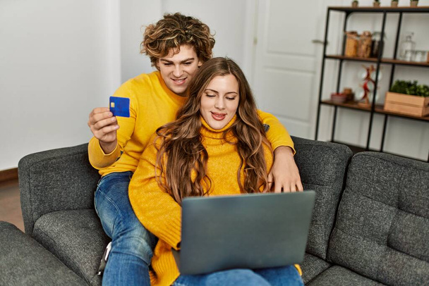 Νεαρό καυκάσιο ζευγάρι που χρησιμοποιεί φορητό υπολογιστή και πιστωτική κάρτα αγκαλιάζονται στο σπίτι - Φωτογραφία, εικόνα