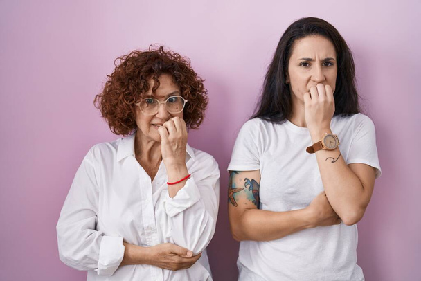 İspanyol anne ve kızı pembe arka plan üzerine günlük beyaz t-shirt giyiyorlar. Elleri ağızda tırnak yerken gergin ve stresli görünüyorlar. anksiyete.  - Fotoğraf, Görsel