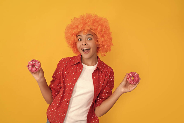 surprised kid in fancy orange wig hair hold sweet glazed donut, surprise. - Фото, изображение