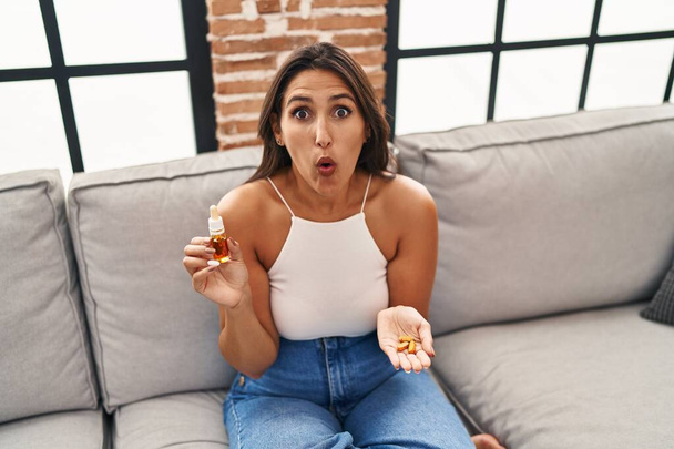 Young hispanic woman holding pills and cbd oil afraid and shocked with surprise and amazed expression, fear and excited face.  - Photo, image