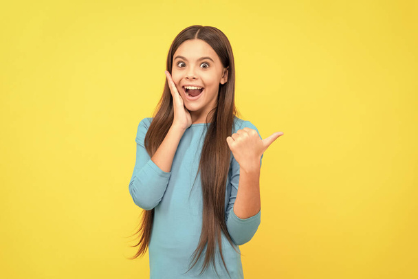amazed teen girl with long hair pointing finger on copy space on yellow background, expressive. - Photo, image