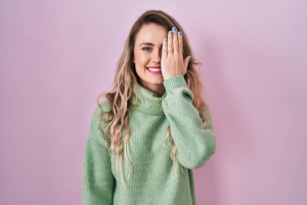 Young caucasian woman standing over pink background covering one eye with hand, confident smile on face and surprise emotion.  - Foto, imagen