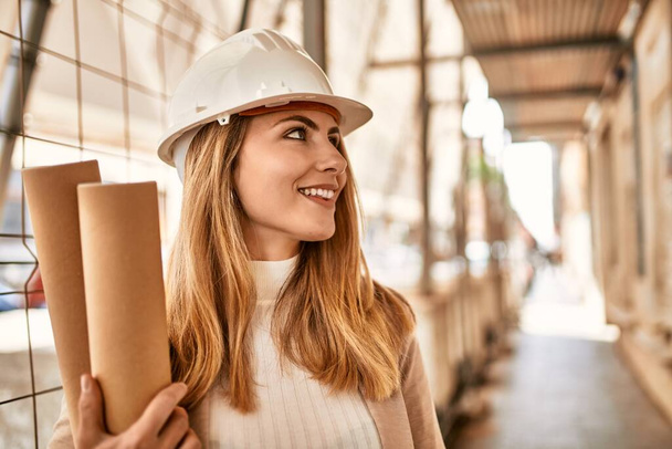 Young blonde woman wearing hardhat holding blueprints at street - Foto, immagini