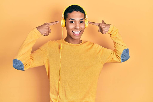 Young african american guy listening to music using headphones smiling pointing to head with both hands finger, great idea or thought, good memory  - Фото, изображение